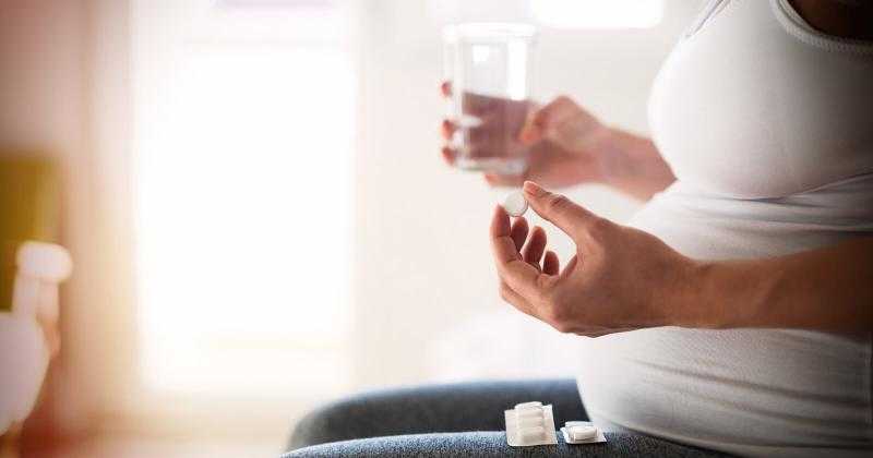 Беременная держит в одной руке таблетку, а в другой - стакан с водой