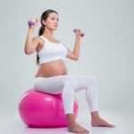 Беременная на фитболе выполняет упражнение с гантелями