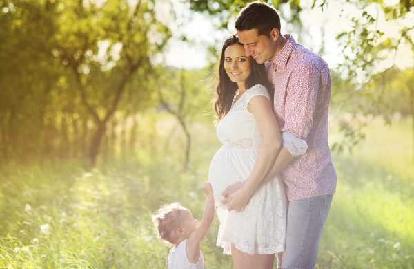 Беременная с мужем и ребёнком стоят в обнимку на фоне деревьев
