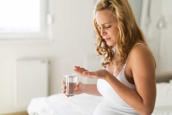 Беременная женщина держит в одной руке таблетку, в другой — стакан воды