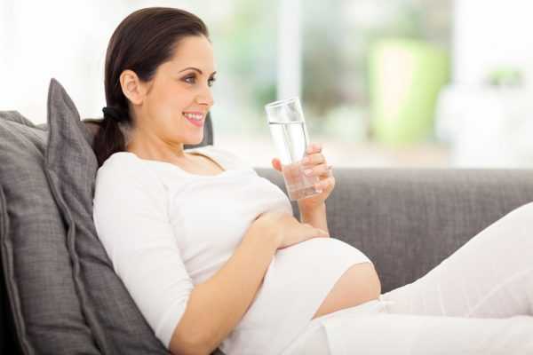 Беременная женщина полулежит на диване, держа в руке стакан с водой