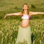 Беременная женщина стоит, раскинув руки, на лугу в солнечную погоду