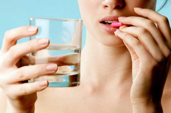 Девушка держит таблетку около рта пальцами одной руки, а другой рукой — стакан с водой