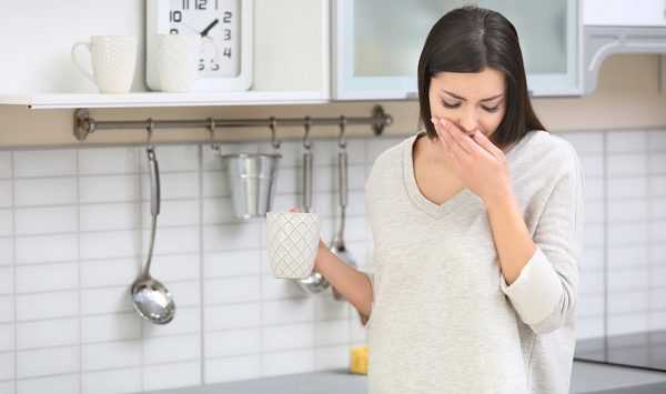 Беременная женщина стоит на кухне и закрывает рот рукой из-за отрыжки
