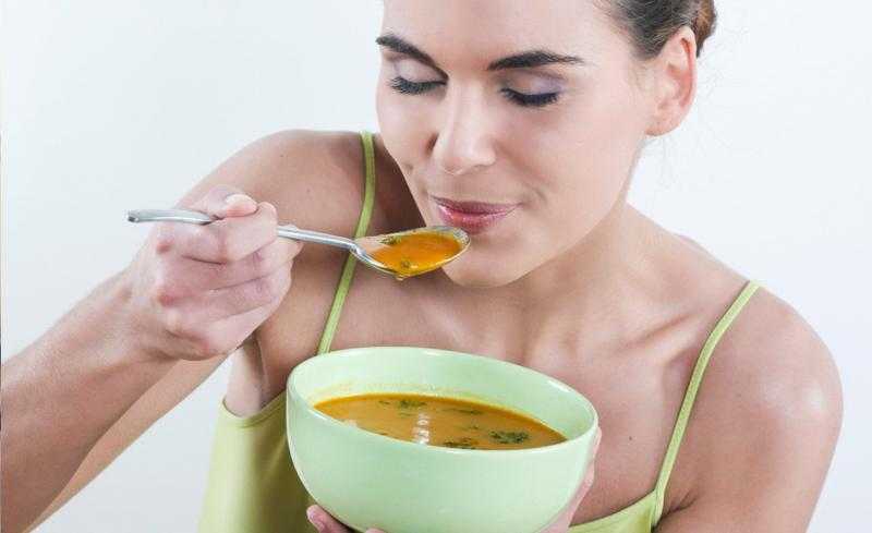 Женщина, прикрыв глаза, держит перед собой тарелку с супом, а в другой руке ложку около рта.