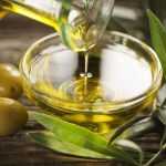 Оливковое масло из бутылки наливают в миску, рядом лежат оливки