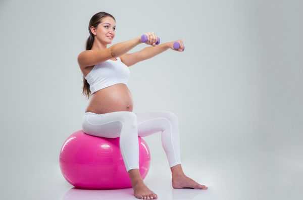 Беременная сидит на мяче и держит гантели в руках, вытянутых вперёд