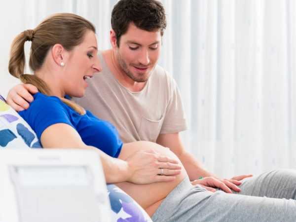 Беременная женщина полулежит и держит руками оголённый живот, рядом с ней сидит муж